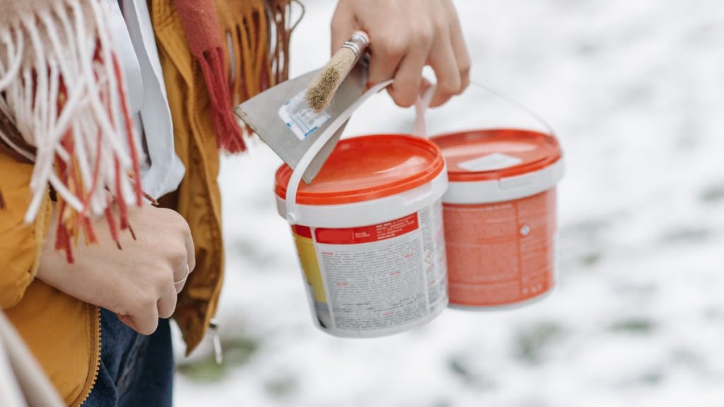 A woman in a yellow jacket holds paint buckets and a brush on a snow-covered day outdoors.