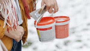 A woman in a yellow jacket holds paint buckets and a brush on a snow-covered day outdoors.