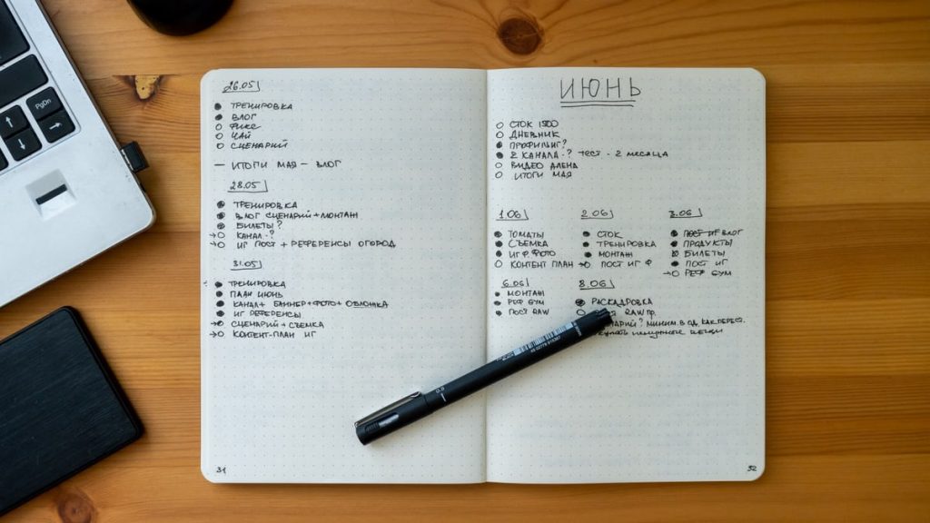 An open notebook with handwritten notes resting on a wooden desk beside a laptop and pen.